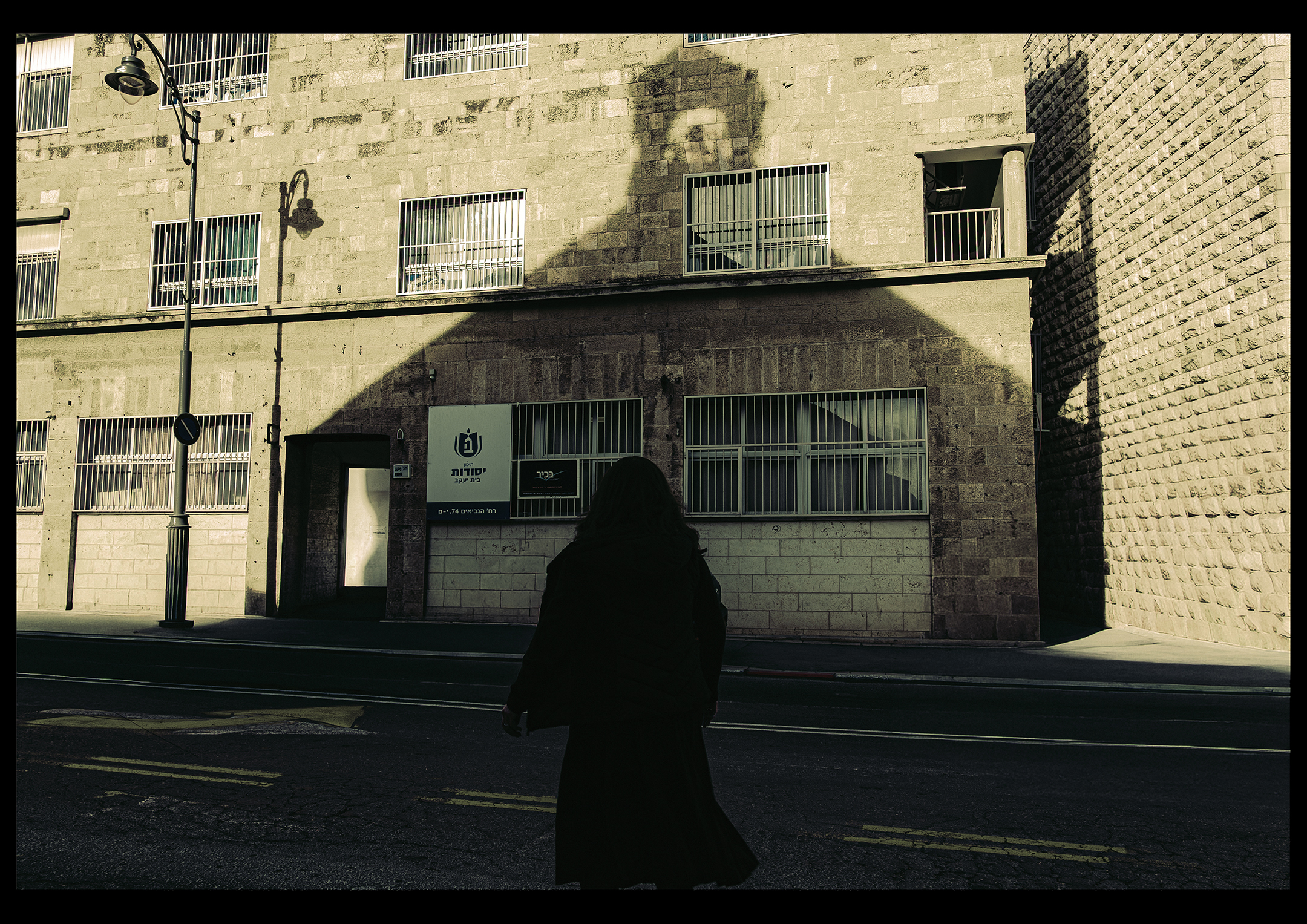 A silhouetted woman crossing a Jerusalem street beneath a large architectural shadow